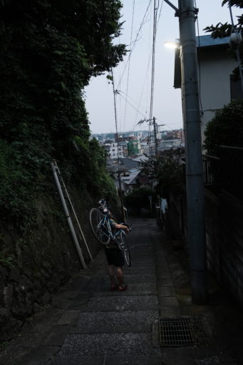 日暮れ時、コンクリートの階段がついた急な坂道を、自転車を肩に担いで下る友達の写真