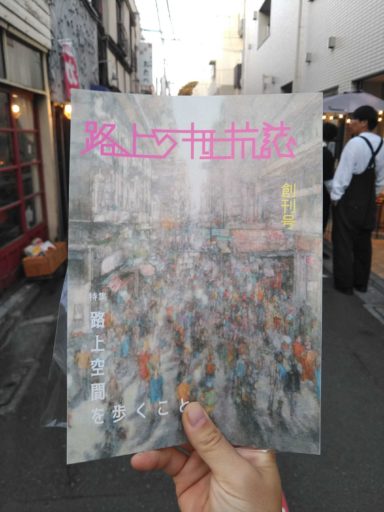 高円寺の街角で『路上の抵抗誌』創刊号を手に持って撮影した写真。沢山の人が行き交う路上を描いた表紙イラストが(スケールはやや異なるが)現実の路地の風景にちょうど重なる格好になっている。