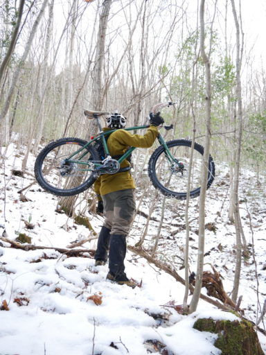 雪の積もった里山の林で自転車を肩に担いで立っている友人の写真