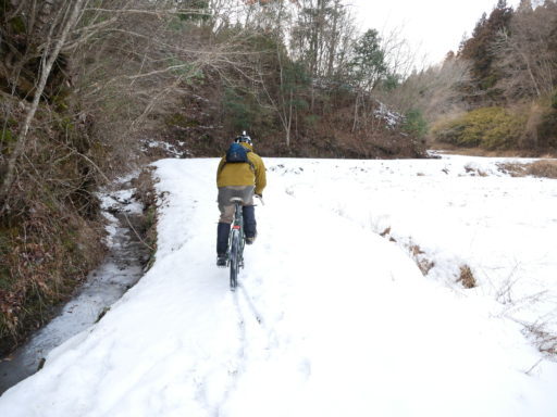 雪の積もった田んぼか畑のへりを自転車で進む友人の姿を撮った写真