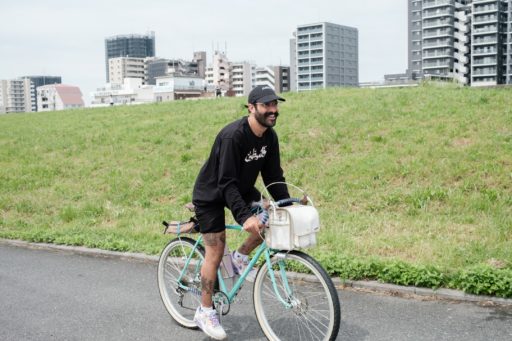 笑顔で自転車を立ち漕ぎしているベトさん。服装は黒いキャップに黒の長袖のTシャツ、あと黒い腿丈のハーフパンツ。腿からふくらはぎにかけてタトゥーが見える。自転車は水色のフレームにドロップハンドル、ランドナースタイルの白いフロントバッグが付いている。向こうに荒川沿いのビル群が見える。