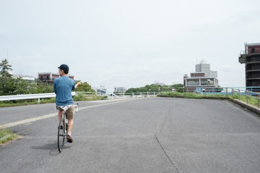 薄曇りの青空の下、両手を離して自転車に乗る筆者。服装はキャップに青い長袖Tシャツ、ベージュのハーフパンツ。腰にクーフィーヤ（パレスチナの伝統的なスカーフ）を巻いている。
