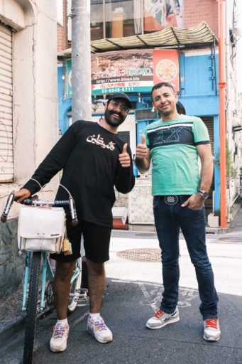 お店の前でベトさんとマンスールさんが親指を立ててこちらを向いている。ベトさんは黒の短パンに胸元にアラビア語で「パレスチナ」とプリントされた黒い長袖Tシャツを着て、横にある自転車を手で支えている。足元は差し色に紫と黄色が入った白いスニーカー。マンスールさんの身長は190cm以上で、服装は緑色のTシャツにジーンズ、赤い靴紐のグレーのスニーカー。ベトさんはマンスールさんより少し背が低い。