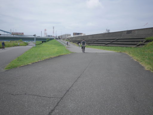 曇り空の下の荒川サイクリングロード。ゆるい下り坂を自転車に乗ったHideさんがこちらに向かってきている。
