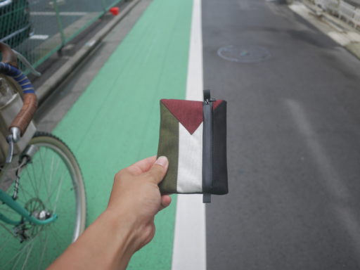 緑色の自転車レーンと黒の通常の道路の間に白い境界線が引かれており、そこにパレスチナ国旗柄のポーチを手に持って重ねている様子。ポーチの柄が緑・白・黒という配色になっており、ちょうど道路の配色と同じになっている。