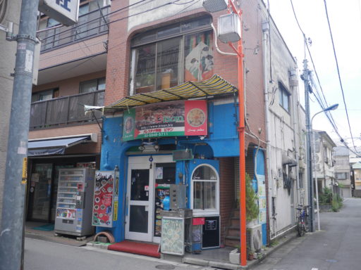 「ビサン」の店構え。レンガ造りの古い2階建てのビルの1階が店舗になっており、店舗の部分は壁が青く、入口のドアが白く塗られている。入口の上に「パレスチナ料理　ビサン」と書かれたパレスチナ国旗の配色の看板。周囲は閑静な住宅街。