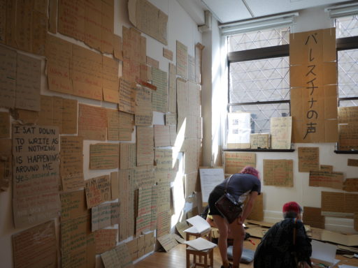 手書きのメッセージが書かれたダンボールで壁が埋め尽くされた部屋。強い西日が差し込んでいる。2人の人がしゃがんで作品を眺めている。奥に一際大きなボードで「パレスチナの声」というタイトルが掲示されている。